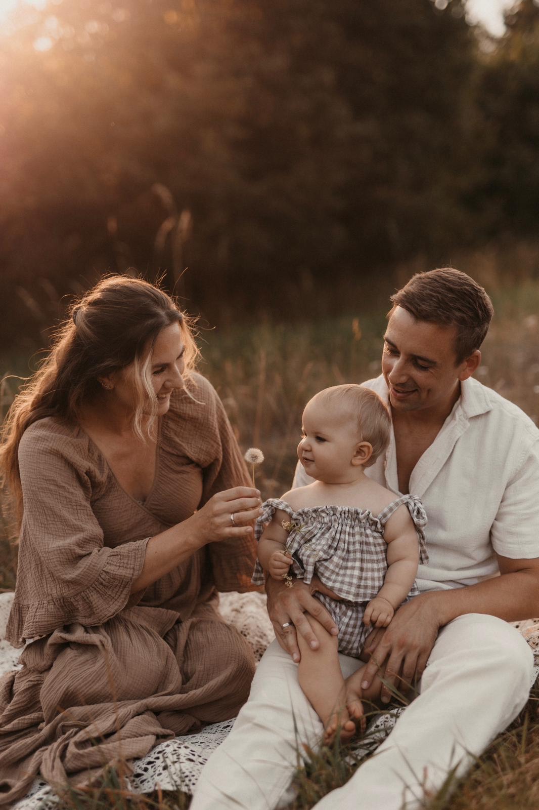 Eltern sitzen mit ihrem Baby auf einer Decke, die Mama hält eine Pusteblume hin