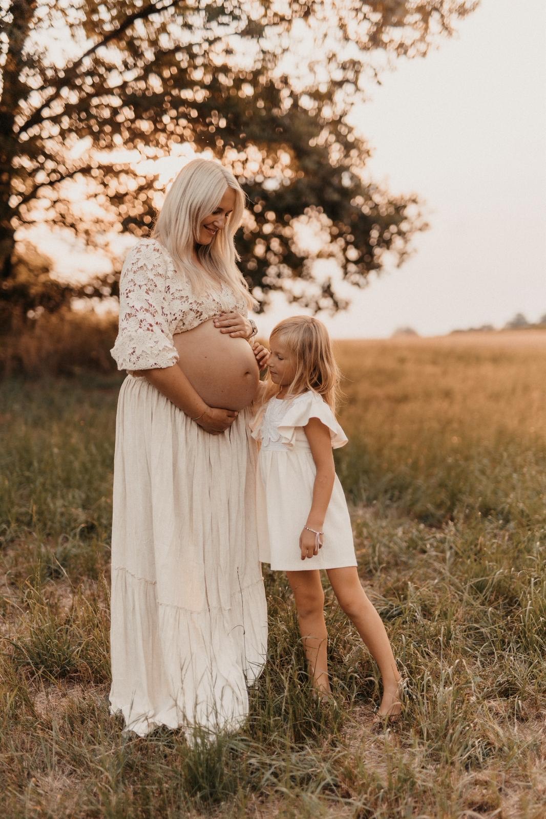 Schwangere Mama im cremefarbenen Kleid, ihre Tochter lehnt sich an den Babybauch