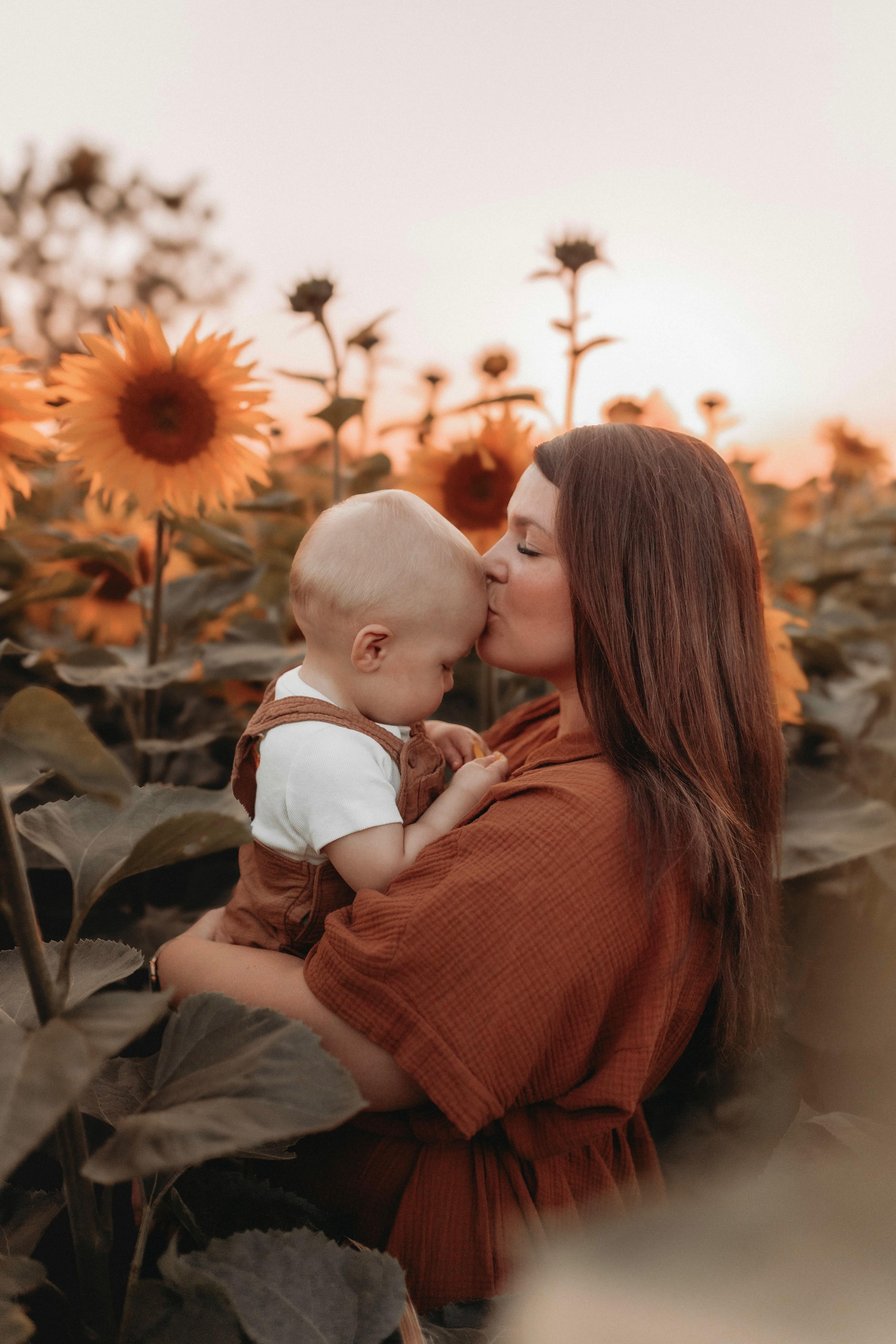 Mutter küsst ihr Baby zärtlich im Sonnenblumenfeld bei Sonnenuntergang – Familienfotografie von Simple Moments